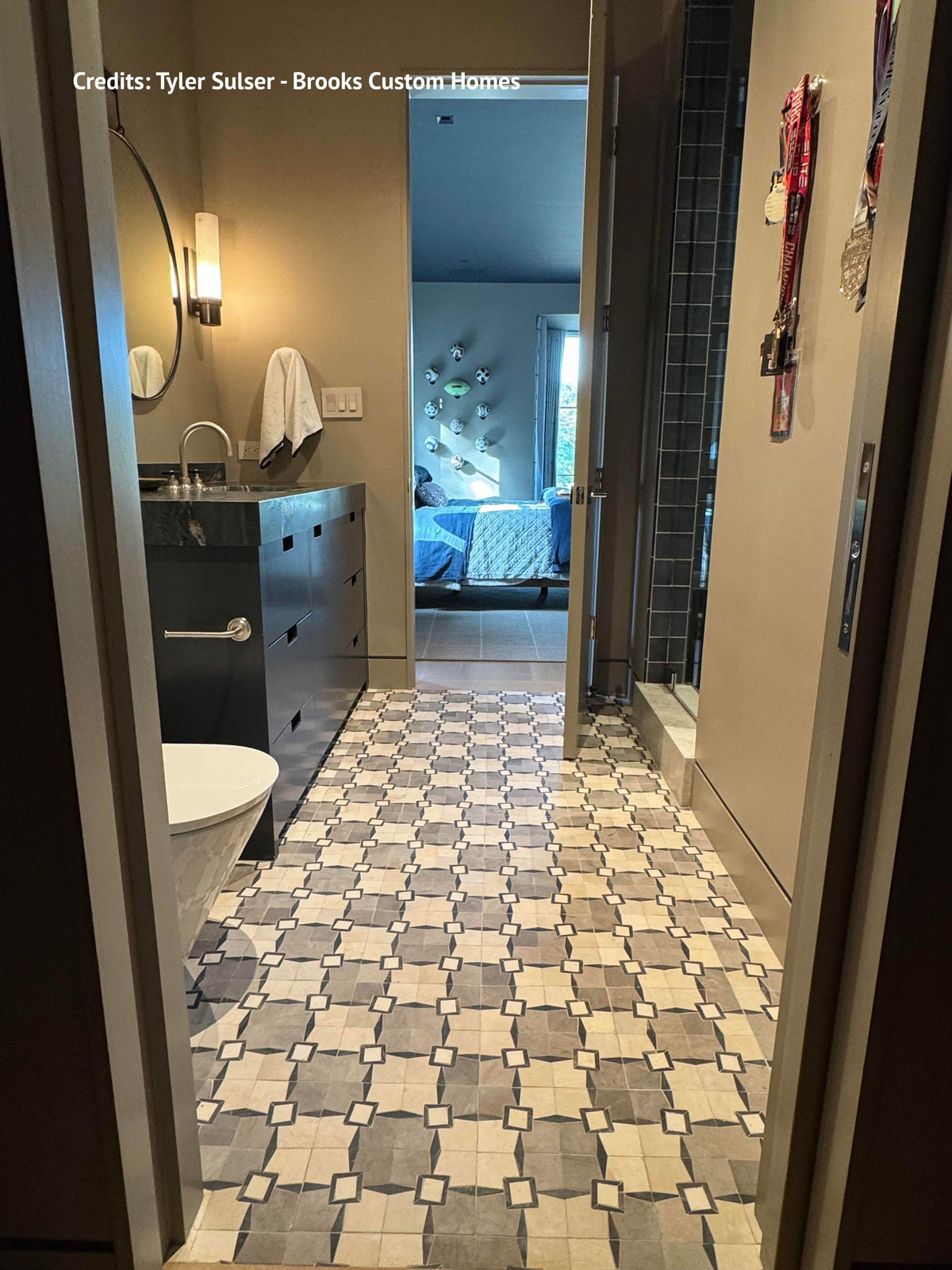 A bathroom interior with a patterned gray and white tile floor, a dark vanity with a sink and mirror, a toilet, and an open doorway leading to a bedroom. Text at the top reads 'Credits: Tyler Sulser - Brooks Custom Homes'.