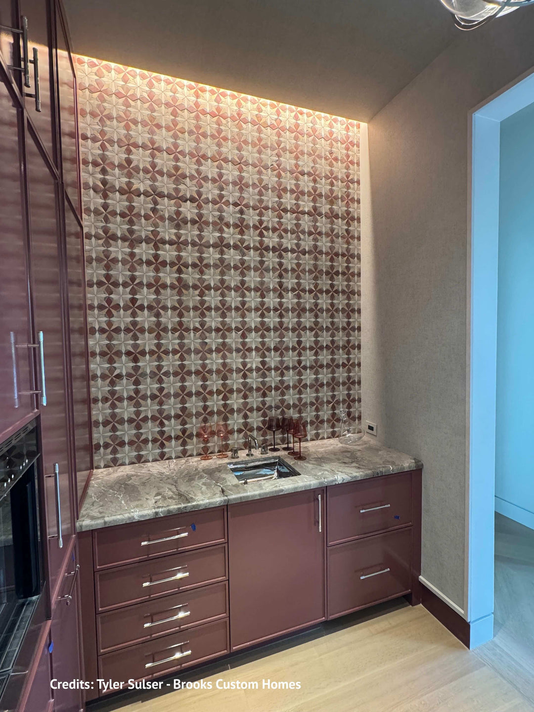 A custom kitchenette corner featuring a patterned brown and beige tile backsplash, a gray marble countertop with an integrated sink, and reddish-brown cabinetry with silver handles.