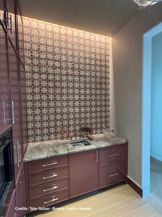 A custom kitchenette corner featuring a patterned brown and beige tile backsplash, a gray marble countertop with an integrated sink, and reddish-brown cabinetry with silver handles.