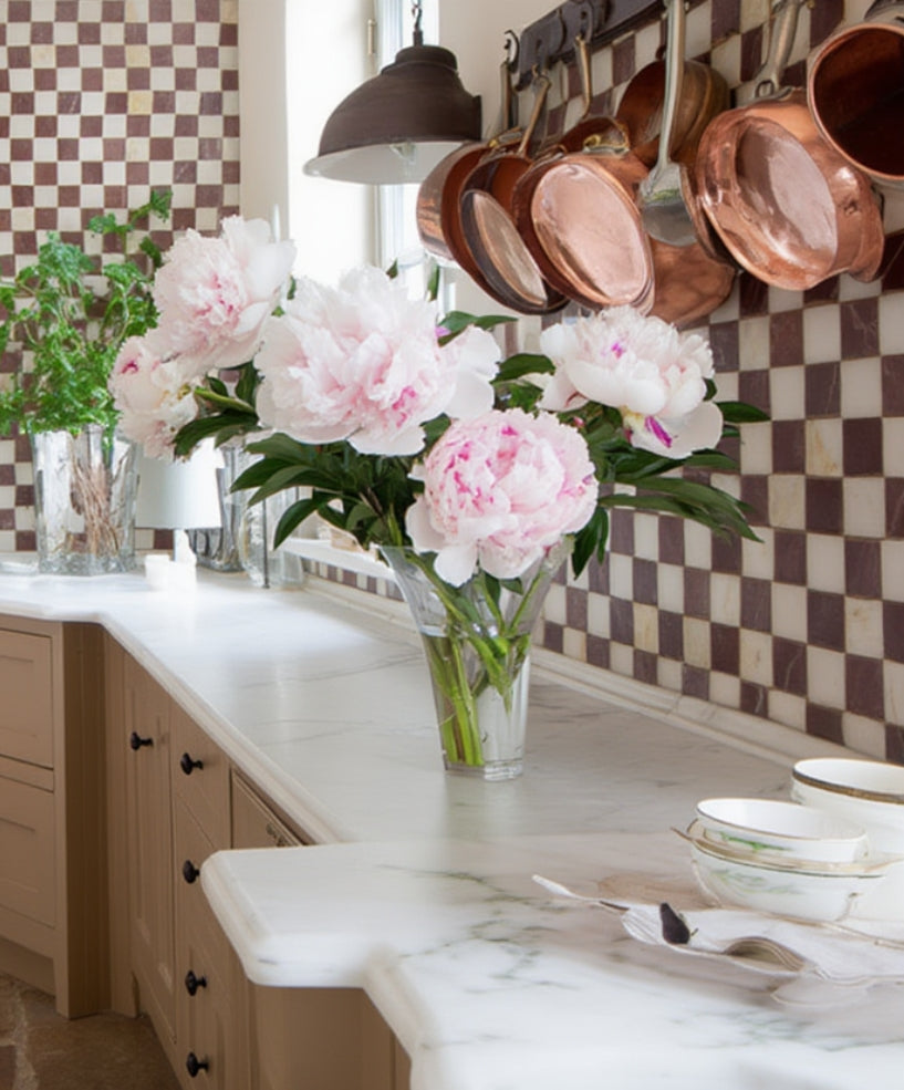 French-style kitchen with marble countertop, burduny-and-white checkered backsplash, copper pots, and pink flowers in a vase.