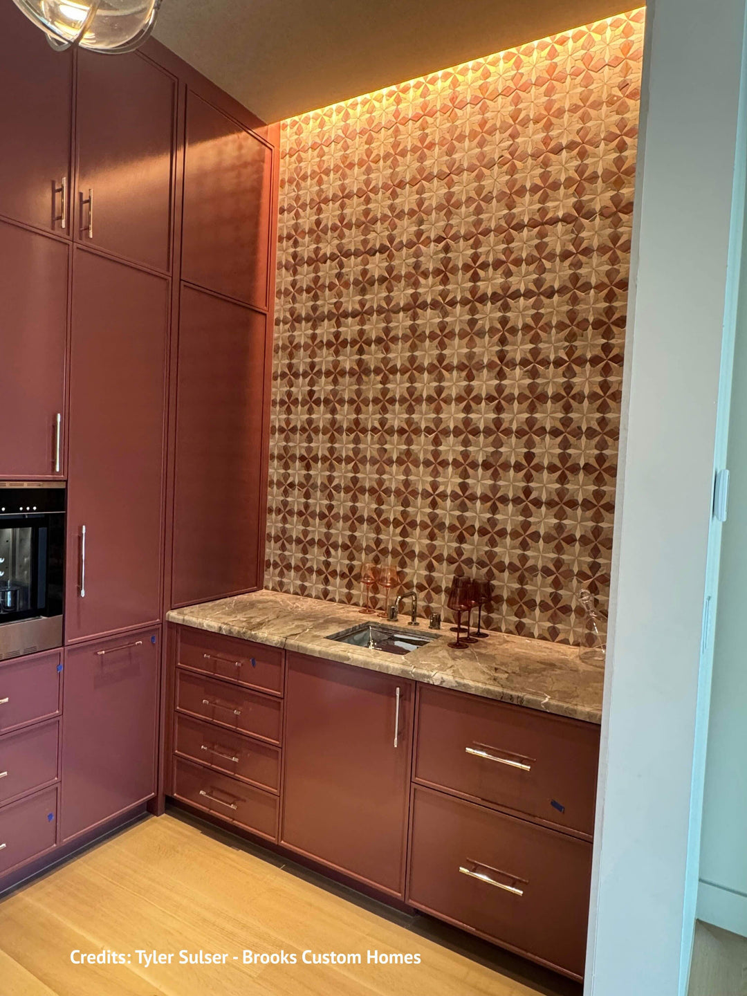 A custom kitchen interior featuring deep pinkish-brown cabinetry, a patterned geometric tile backsplash in warm brown and gold tones, a marble countertop, and light wood flooring.