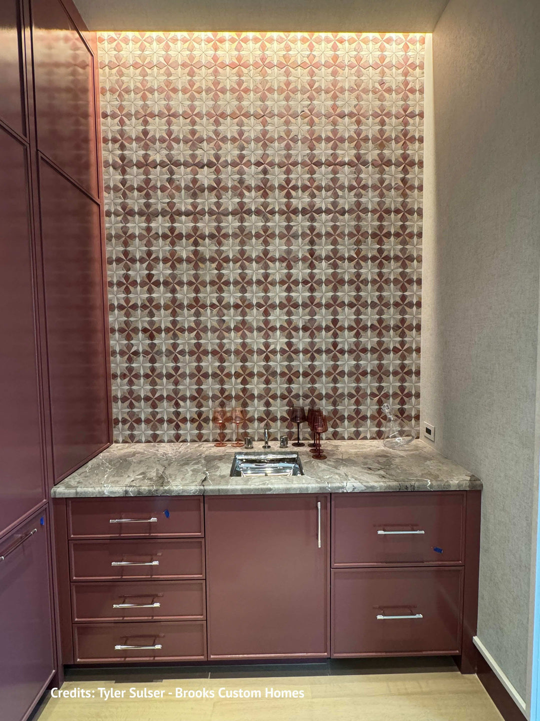 A kitchen cabinetry and backsplash setup featuring a patterned brown and beige tile backsplash, a gray marble-look countertop, and dark reddish-brown lower cabinets with silver handles.