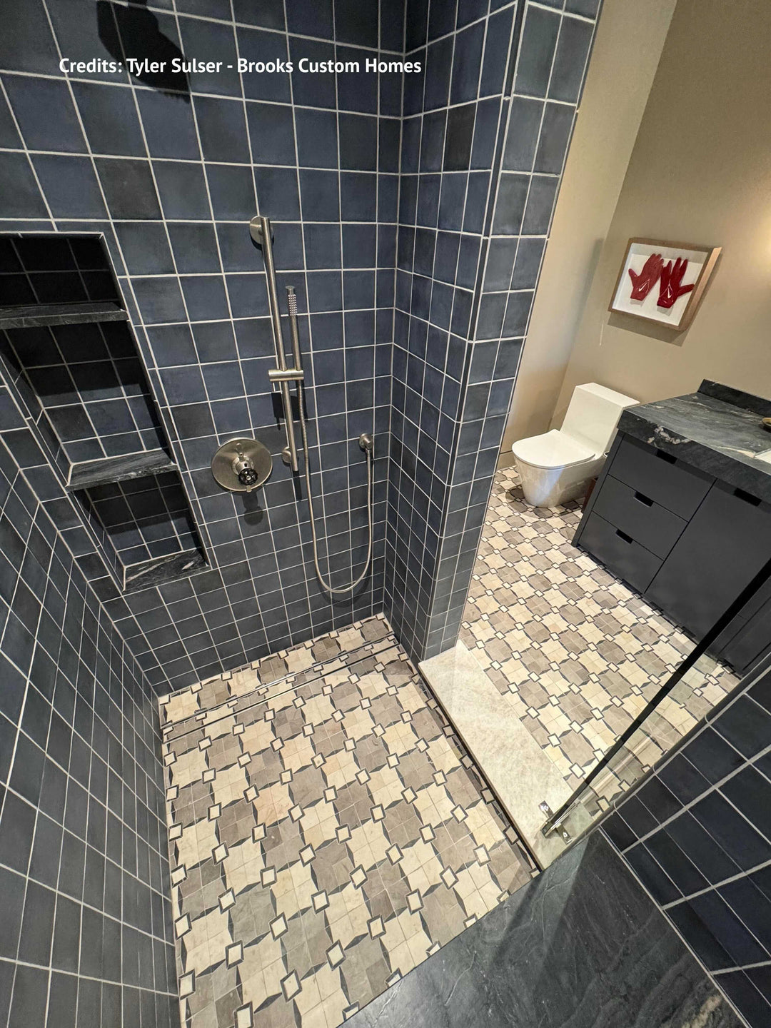 A modern bathroom shower area with dark blue-gray square wall tiles, a patterned beige and gray floor, a built-in niche, and a glass shower door. A white toilet and dark vanity are visible in the adjacent bathroom space.